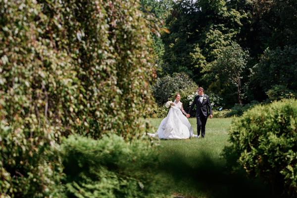 نمای جلوی زوج متاهل خوشبخت که در روز عروسی در فصل تابستان با دستانشان به یکدیگر نگاه می کنند و در حال قدم زدن در پارک شگفت انگیز هستند