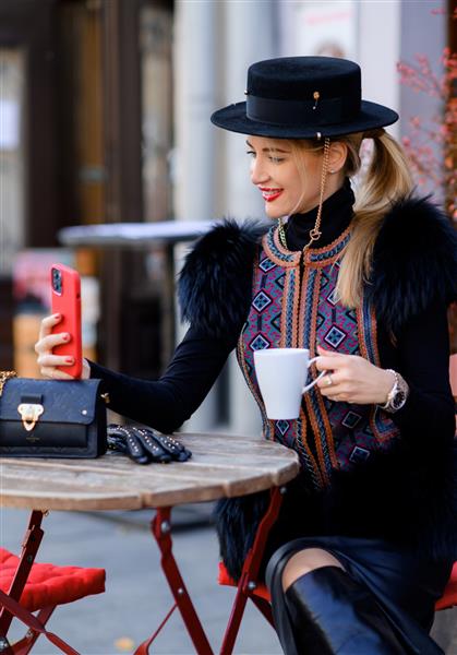 نمای نزدیک از زن جذاب با کلاه سیاه با زنجیر با لباس جلیقه شیک با خز و گلدوزی که با تلفن همراه پشت میز نشسته و در حال نوشیدن قهوه در کافه تریا عکس سلفی می گیرد