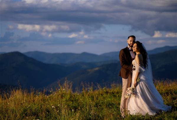 تازه عروس هایی که به غروب آفتاب نگاه می کنند و روی تپه ژست گرفته اند