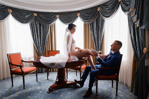 نمای کناری عروس جنسی با لباس زیر سفید و چادر بلند روی موها روی میز نشسته و به داماد نگاه می کند که روی صندلی نشسته و کفش عروسی را در اتاق هتل در دست گرفته است