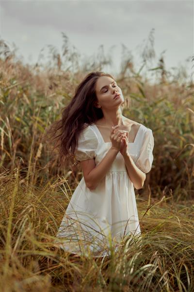 زن جوان زیبا با لباس تابستانی سفید در چمن های بلند در زمین روستایی نشسته است پرتره دختر عاشقانه در غروب آفتاب گرم زن زیبای طبیعی