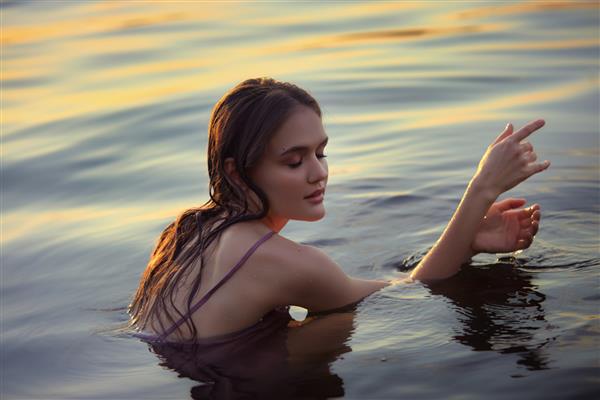 زن جوان زیبا در آب دریاچه با لباس تابستانی در غروب آفتاب پرتره یک دختر خیس عاشقانه در غروب آفتاب گرم زیبایی طبیعی یک زن