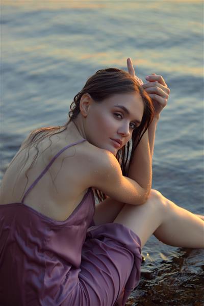 زن جوان زیبا نشسته در ساحل دریاچه با لباس تابستانی در غروب آفتاب پرتره یک دختر خیس عاشقانه در غروب آفتاب گرم زیبایی طبیعی یک زن
