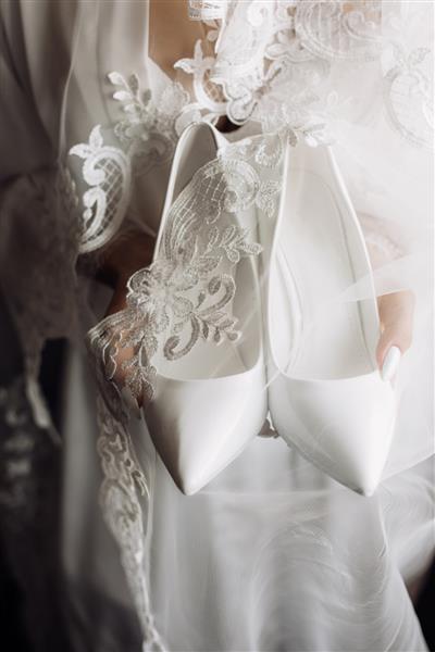 کفش تشریفاتی عروسی سفید در دستان عروس با لباس شب ابریشمی توری