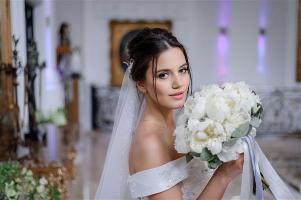 عروس زیبای سبزه قفقازی دسته گل گل صد تومانی سفید را در دست گرفته و در داخل خانه مستقیم به نظر می رسد