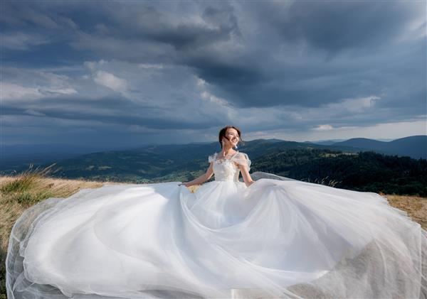 عروس شاد زیبا با لباس عروسی لوکس در روز آفتابی در کوهستان با آسمان ابری
