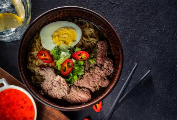 سوپ رشته فرنگی آسیایی با گوشت گاو تخم مرغ فلفل قرمز و گیاهان روی میز تاریک نمای بالا