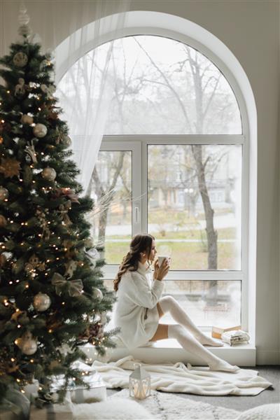 دختر زیبا با یک فنجان چای و چهارخانه کنار درخت کریسمس نشسته است
