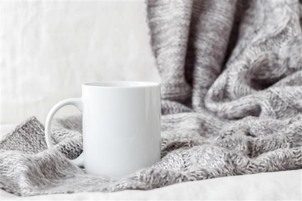 ماکت لیوان قهوه سفید روی تخت با پتوی خاکستری
