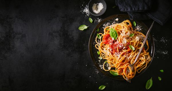 ماکارونی اسپاگتی کلاسیک ایتالیایی خوشمزه و اشتها آور با سس گوجه فرنگی پنیر پارمزان و ریحان در بشقاب روی میز تاریک نمای از بالا افقی