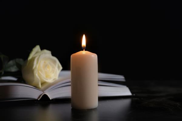 شمع سوزان کتاب و رز سفید روی میز در تاریکی فضایی برای متن نماد تشییع جنازه