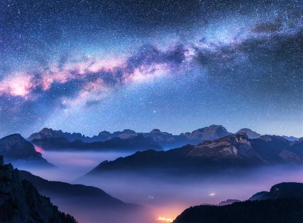راه شیری بر فراز کوه ها در مه در شب در پاییز منظره ای با دره کوه آلپ ابرهای کم ارتفاع آسمان پرستاره بنفش با کهکشان راه شیری روشنایی شهر هوایی Passo Giau دولومیت ایتالیا فضا