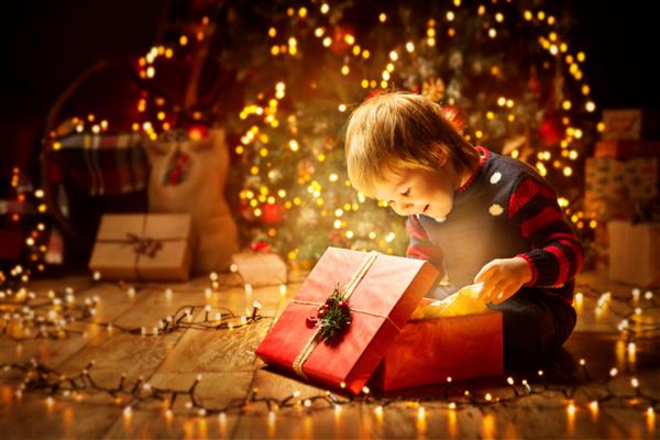 هدیه باز کریسمس برای کودک پسر بچه شادی که به دنبال نور جادویی در جعبه است کودکی که جلوی درخت کریسمس نشسته است