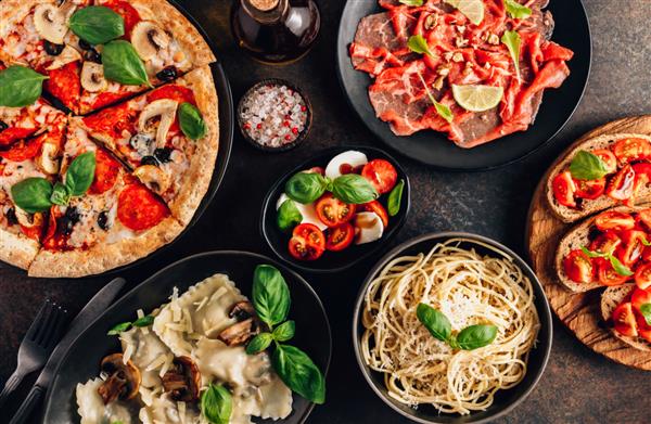 میز کامل غذاهای ایتالیایی در بشقاب پیتزا پاستا راویولی کارپاچیو سالاد کاپرز و بروشتا گوجه فرنگی در زمینه مشکی نمای بالا