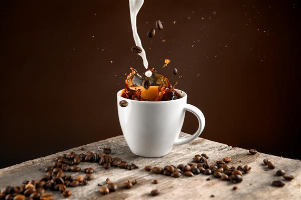 عکس استودیویی اسپلش از دانه‌های قهوه و شیر که داخل فنجان سفید پر از قهوه می‌افتند