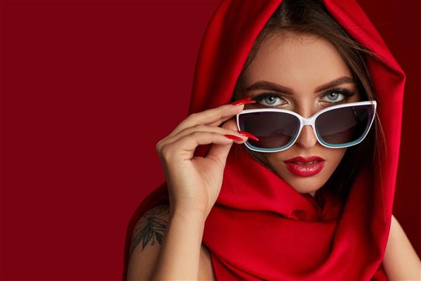 پرتره دختر زیبای سبزه با آرایش مجلل با عینک آفتابی سفید و روسری قرمز در پس زمینه قرمز زن به دوربین نگاه می کند