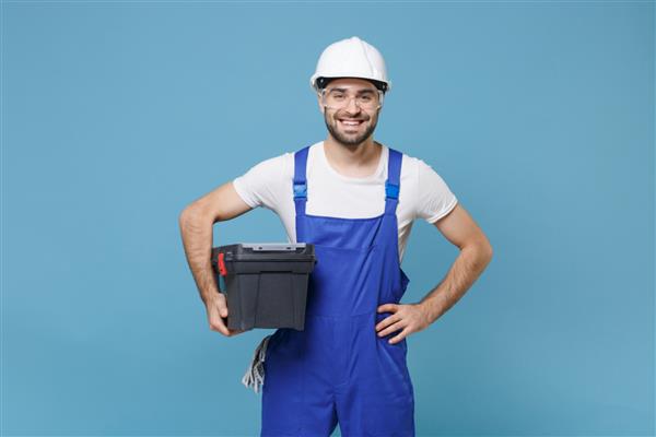 مرد خندان با کلاه ایمنی کلاه ایمنی که با عینک جعبه ابزار جدا شده روی پس‌زمینه آبی قرار دارد لوازم جانبی ابزار برای بازسازی اتاق آپارتمان مفهوم خانه تعمیر ایستاده با بازوهای کیمبو