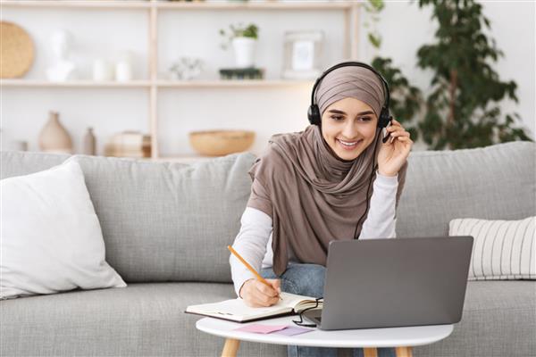 آموزش آنلاین برای زنان مسلمان دختر عربی شاد با روسری و هدست در حال مطالعه با لپ تاپ در خانه یادداشت برداری در حین تماشای وبینار