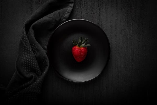 عکاسی از غذای تیره - یک توت فرنگی روی یک بشقاب سیاه با پس زمینه تیره و لوازم