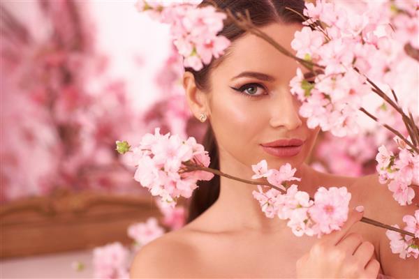 پرتره صورت بهاری مد نزدیک از زن جوان زیبای قفقازی با موهای سبزه در دم اسبی و آرایشی عالی که از میان درختان با گل های صورتی در شکوفه نگاه می کند