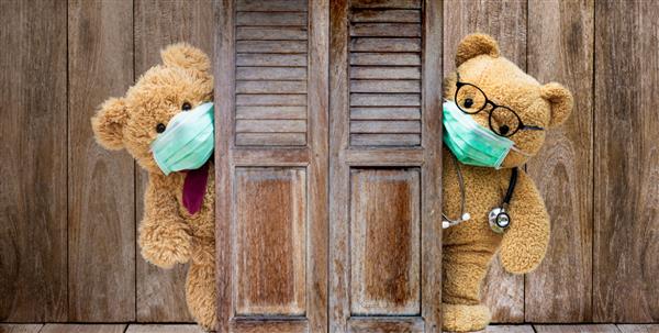 خرس عروسکی مریض قهوه ای ماسک پزشکی محافظ می پوشد و پزشک با گوشی پزشکی پشت در یا پنجره چوبی قدیمی ایستاده است در خانه بمانید قرنطینه پیشگیری از همه گیری کرونا ویروس مفهوم کووید-19