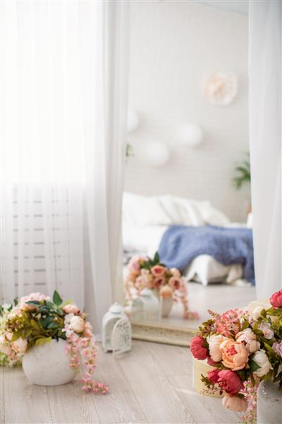 آینه بلند در قاب شیک پوشیده شده با پارچه توری سفید در کنار گلدان های گلدار انعکاس اتاق خواب روشن تخت بزرگ با کتانی سفید و پتوهای بافتنی مفهوم آسایش و تفریح در خانه