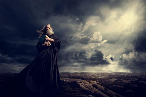 راهب که کتاب مقدس را در دست دارد و به نور آسمان نگاه می کند کشیش پیر با لباس سیاه در کوه های طوفان