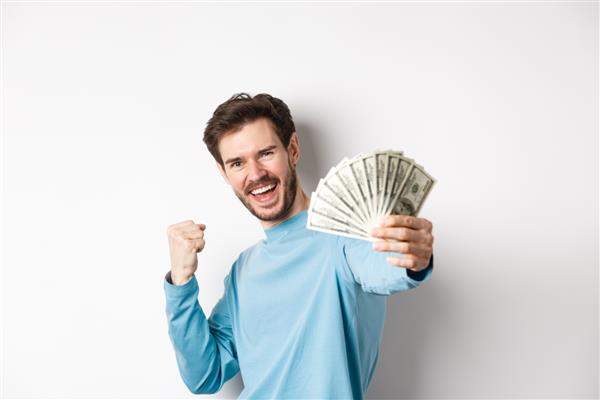 مرد خوشبخت قفقازی دستش را با پول به دلار دراز می کند بله می گوید و درآمد را جشن می گیرد جایزه نقدی دریافت کرد روی پس زمینه سفید ایستاده بود