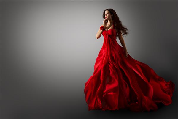 زن مد لباس قرمز مدل زیبایی در حال رقصیدن با لباس شب بلند که روی موهای مشکی باد در حال پرواز در هوا بر روی پس زمینه خاکستری است