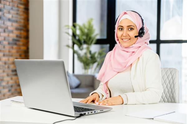 پرتره تجاری زن مسلمان با حجاب هدست بی سیم با استفاده از لپ تاپ برای کار در دفتر معاصر تاجر خاورمیانه ای به دوربین نگاه می کند و پشت میز نشسته لبخند می زند