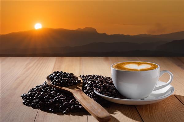 فنجان قهوه لاته سفید با شکل قلب و دانه‌های قهوه روی میز چوبی غروب تار در پس‌زمینه کوه قاشق چوبی نوشیدنی صبحگاهی مفهوم غذا و نوشیدنی