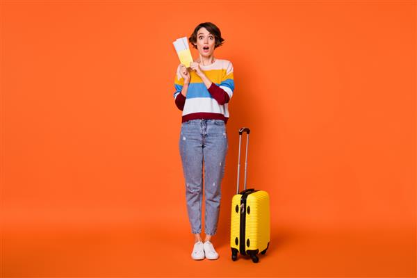 عکس بانوی جوان جذاب و هیجان‌زده با چمدان‌های پیراهن کش راه راه لباس پوشیده در حالی که بلیط در دست دارد پس زمینه نارنجی جدا شده