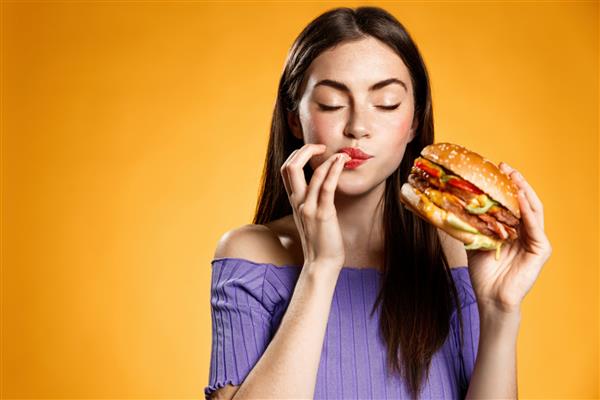 زن در حال خوردن چیزبرگر با رضایت دختر از غذای خوشمزه همبرگر لذت می برد لقمه خوشمزه همبرگر را لیس می زند در حالی که گرسنه است سفارش تحویل فست فود می دهد روی پس زمینه نارنجی ایستاده است