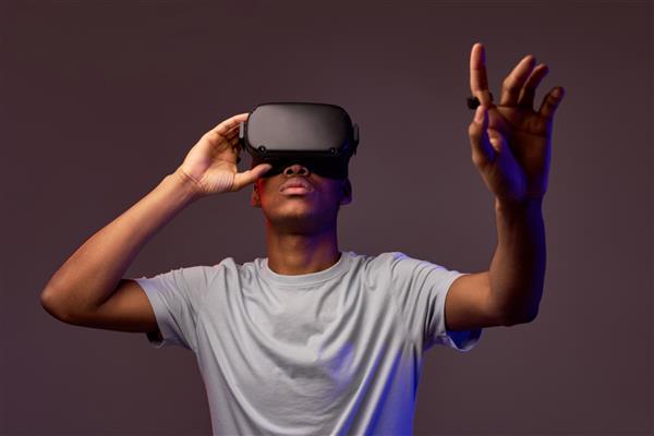 پرتره پسر نوجوان آفریقایی با کلاه VR به فضای کپی نگاه می کند در استودیو جدا شده است پسری دستش را به جلو دراز کرده است گیمر در حال انجام بازی های VR در پس زمینه رنگارنگ بازی VR