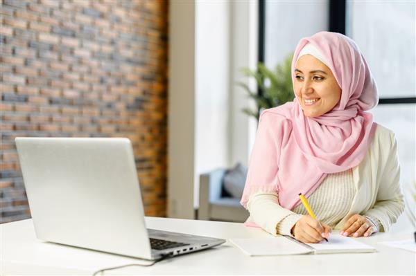 تاجر مسلمان خندان که پشت میز در فضای اداری مدرن نشسته یادداشت برداری می کند دختر دانشجوی مسلمان جوان با حجاب و لباس راحتی هوشمند از صفحه لپ تاپ روی دفترچه یادداشت می نویسد