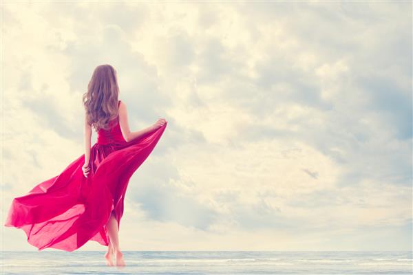 زن از دریای خورشید در ساحل لذت می برد نمای پشتی دختر در لباس پرنده قرمز در باد تعطیلات تعطیلات مبارک سبک زندگی رفاهی