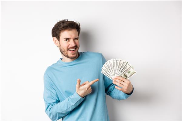 مرد ثروتمند خوش تیپ در حال چشمک زدن به دوربین نشان دادن درآمد نشان دادن انگشت به سمت پول به دست آمده و لبخند زدن ایستادن روی پس زمینه سفید