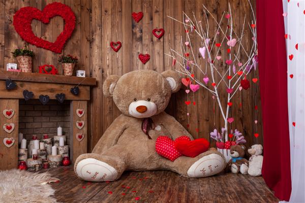 پس زمینه زیبا برای عکسبرداری روز ولنتاین با یک خرس عروسکی بزرگ یک شومینه چوبی با شمع ها و گل ها و یک درخت با قلب های قرمز و صورتی خلق و خوی رمانتیک برای تعطیلات عاشقان