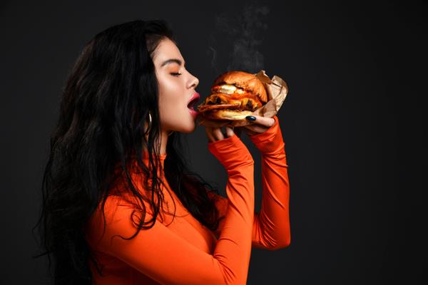 پرتره زن سبزه سکسی داغ با لب های پف کرده در لباس قرمز در حال گاز گرفتن همبرگر بزرگ دستی تازه خوشمزه با بخار روی پس زمینه سیاه مفهوم فست فود و رژیم غذایی