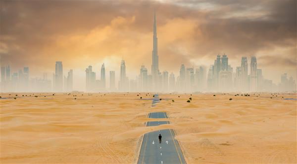 نمایی از بالا نمای هوایی خیره کننده از یک فرد ناشناس که در جاده ای متروک پوشیده از تپه های شنی با افق دبی در پس زمینه راه می رود دبی امارات متحده عربی