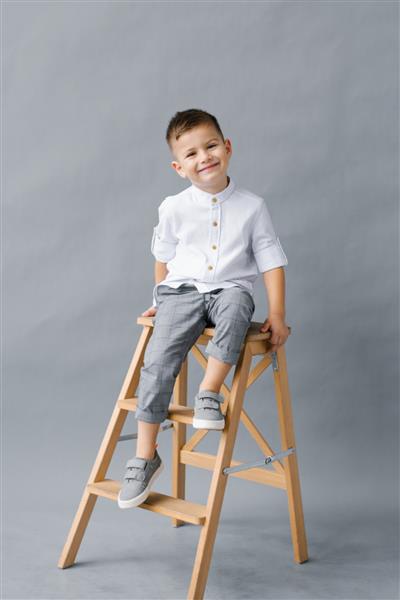 پسر زیبا و شیک روی یک پله چوبی روی پس زمینه خاکستری نشسته و لبخند می زند