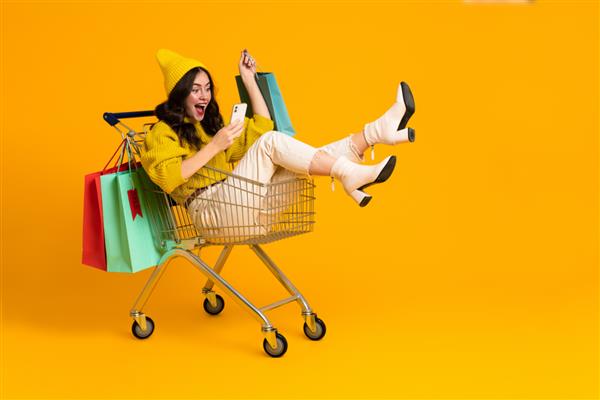 زن هیجان‌زده سفیدپوست در حال خندیدن و استفاده از تلفن همراه در سبد خرید جدا شده روی پس‌زمینه زرد