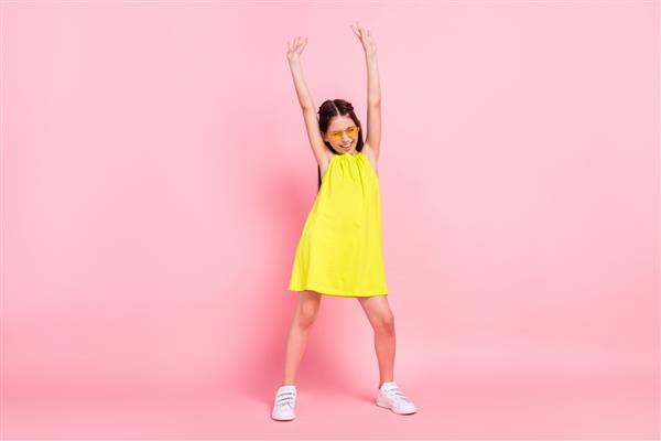 عکس تمام قد از دختر مدرسه ای زیبا و جذاب با لباس زرد عینک در حال رقصیدن با بازوهای بلند شده با پس زمینه صورتی رنگ