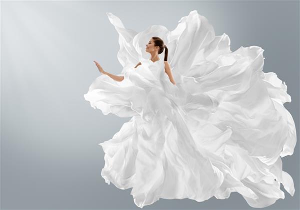 مدل مد در لباس سفید خالص خلاقانه به عنوان ابر زنی با لباس ابریشمی بلند با پارچه ابریشمی که روی باد بر روی پس زمینه خاکستری روشن پرواز می کند هنر فانتزی دختر رقصنده