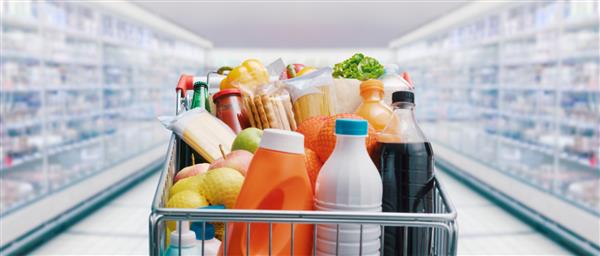 سبد خرید پر از غذا و نوشیدنی و قفسه‌های سوپرمارکت در پس‌زمینه مفهوم خرید مواد غذایی