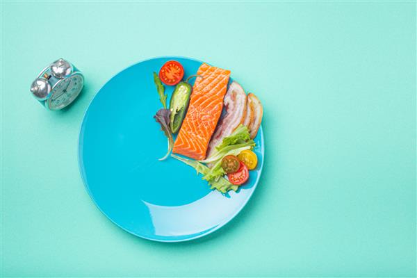 ناشتا متناوب مفهوم رژیم غذایی کم کربوهیدرات با چربی بالا غذای سالم ماهی سالمون گوشت بیکن سبزیجات و سالاد در بشقاب آبی و زنگ ساعت در نمای بالای پس زمینه آبی فضایی برای متن