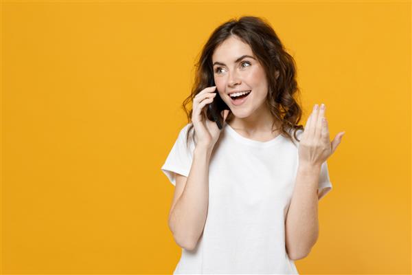 زن جوان خندان و شاد قفقازی با تی شرت ساده و معمولی سفید و با تلفن همراه صحبت می کند و مکالمه دلپذیری را انجام می دهد که با دست روی پس زمینه زرد نارنجی پرتره استودیویی است