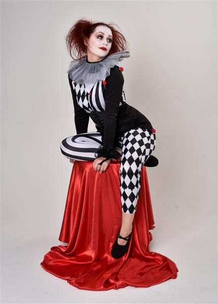 پرتره تمام قد دختری با موهای قرمز با لباس دلقک سیاه و سفید شخصیت سیرک تئاتر نشستن روی صندلی جدا شده روی پس‌زمینه استودیو
