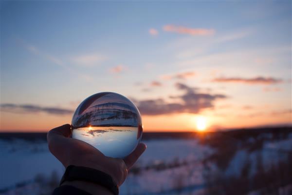 منظره زیبا از طریق لنز توپ کریستالی در افق عصر برفی در غروب زمستان زمستان بازتاب ابرها در کره کره زمین را کف دست در فضای باز نگه می دارد