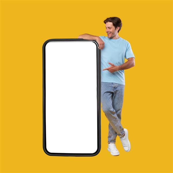 تبلیغات اپلیکیشن موبایل تمام قد مرد شادی که به صفحه نمایش گوشی هوشمند خالی بزرگ و سفید و بزرگ که روی پس‌زمینه استودیو نارنجی ایستاده است اشاره می‌کند این را بررسی کنید مدل نمایشگر تلفن همراه